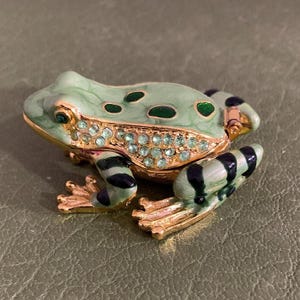 Pode incluir: Estatueta decorativa de sapo em tons de verde e dourado. O sapo tem um corpo verde claro com manchas verdes escuras e listras pretas nas pernas. A barriga do sapo é adornada com pequenos cristais transparentes. A estatueta é feita de metal e tem uma tampa articulada.