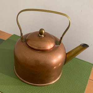 Könnte beinhalten: Ein kupferfarbener Teekessel mit rundem Körper, einem messingfarbenen Griff und einem passenden messingfarbenen Ausguss. Der Kessel verfügt über einen abnehmbaren Deckel mit einem kleinen, abgerundeten messingfarbenen Knopf in der Mitte.
