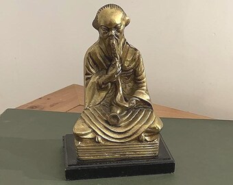 Op de afbeelding: Een messingkleurig beeld van een zittende, oudere figuur met een lange baard en een gewaad. Het beeld is gemonteerd op een zwarte rechthoekige basis. De figuur houdt een klein voorwerp bij de mond.