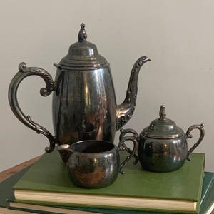 May include: A silver-plated tea set consisting of a tall teapot with a lid, a creamer, and a lidded sugar bowl. Each piece features ornate, curved handles and decorative detailing on the spouts and finials, showcasing a polished metallic finish.