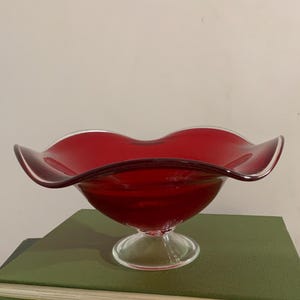 May include: A deep red glass bowl featuring a wide, undulating, ruffled rim and a clear glass pedestal base. The bowl has a smooth, translucent finish and a curved, open design that flares outward from the center to the decorative wavy edge.