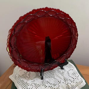 Op de afbeelding: Een robijnrode glazen serveerschaal met een geschulpte rand en ingewikkelde reliëfdetails. De ronde schaal staat op een zwarte standaard en toont zijn levendige kleur en ontwerp. Een decoratief stuk voor serveren of tentoonstellen.