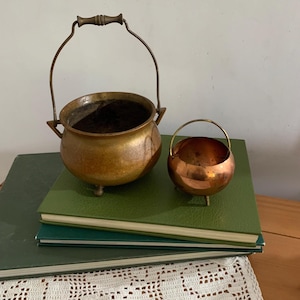 May include: A large brass cauldron with a swing handle and three small feet sits beside a smaller, polished copper pot with a thin wire handle and three feet. Both metal vessels feature rounded bodies and open tops, showcasing a metallic finish.