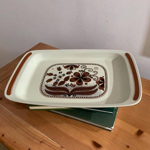 Puede incluir: Bandeja rectangular crema con diseño floral marrón. La bandeja tiene asas marrones y un estampado floral decorativo en el centro. La bandeja está sobre una pila de libros.