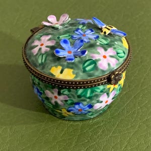 May include: A small, round trinket box with a green base and lid, adorned with painted flowers in pink, blue, and yellow. The lid features a raised butterfly and flower accents. The box has a brass-colored rim and clasp.