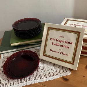 May include: A stack of deep red glass dessert plates and one individual plate featuring a textured rim pattern. The items are accompanied by a white cardboard box labeled Avon 1876 Cape Cod Collection Two Dessert Plates with red and gold borders.