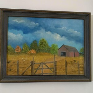 May include: Oil painting of a rural landscape with a red barn, wooden gate, and a fence. The sky is blue with white clouds. The painting is in a dark wooden frame.