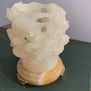 May include: A carved, off-white decorative vase with a dragon motif. The vase sits on a round, amber-colored base with gold-colored accents. The vase has a textured surface and a unique, open design.