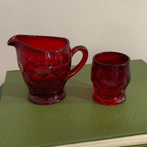 May include: A ruby red glass creamer pitcher and a matching small glass tumbler, both featuring a raised hexagonal honeycomb pattern across their exterior surfaces. The creamer has a curved pouring spout and a side handle, while the tumbler is cylindrical.