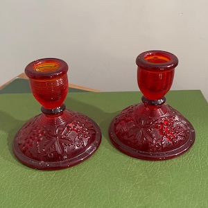 Peut inclure: Deux bougeoirs en verre rouge. Chacun a une base ronde avec un motif de raisin et de feuilles, une courte tige et un sommet en forme de coupe. Les bougeoirs sont en verre rouge.