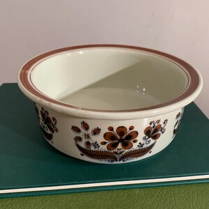Puede incluir: Un cuenco redondo de cerámica blanquecina con borde marrón y estampado floral. El cuenco presenta flores y follaje estilizados en marrón y marrón oscuro en el exterior. El cuenco está sobre una superficie verde.
