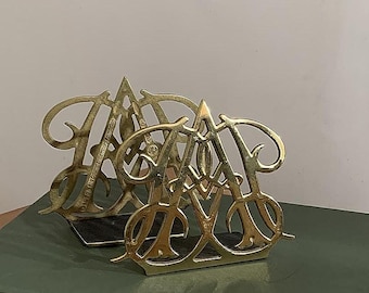 May include: Two gold-colored metal bookends in the shape of the letter "A". The bookends have an ornate, intertwined design and are resting on a green surface. The bookends are likely vintage or antique.