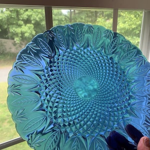 May include: A round, iridescent turquoise glass plate featuring a central diamond-patterned texture surrounded by a border of embossed leaf motifs. The plate has a scalloped edge and exhibits a reflective, semi-translucent finish that catches light.