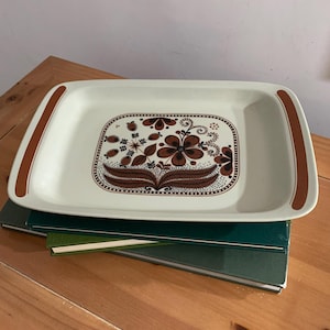 Puede incluir: Bandeja rectangular para servir con base color crema y diseño floral marrón. La bandeja tiene detalles marrones en las asas. El diseño floral presenta flores y hojas marrones. La bandeja está colocada sobre una pila de libros verdes.