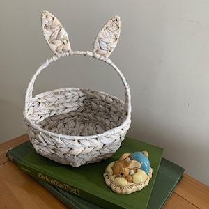 May include: A woven basket with bunny ears attached to the handle. The basket is white and beige. A ceramic figurine of a bunny resting near a blue egg is on a green book.