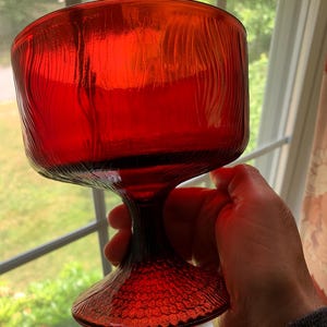May include: A vibrant red glass goblet featuring a textured, vertical ribbed pattern on the bowl and a distinct honeycomb-like embossed design on the circular base. The glass is translucent, allowing light to pass through its deep crimson surface.