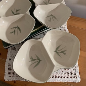 May include: Three off-white ceramic divided serving dishes with a smooth, rounded shape. Each section of the dishes features a delicate, muted green bamboo leaf illustration centered at the bottom. The edges of the bowls are finished with a thin, subtle green trim.