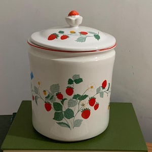 May include: A white ceramic canister with a matching lid featuring a red strawberry knob. The body and lid are decorated with a pattern of red strawberries, green leaves, and small white flowers. A thin red line accents the rim of the lid.