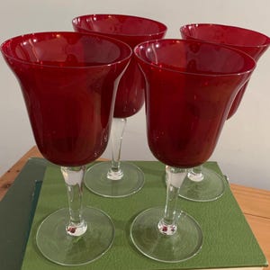 May include: Four ruby red wine glasses with clear stems and circular bases. The glasses have a tulip-shaped bowl. The glasses are arranged on a green surface.