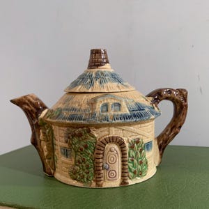 Puede incluir: Tetera de cerámica con forma de cabaña, con tejado de paja y detalles azules. La tetera tiene un asa y un pico marrones, con ventanas y una puerta. Un artículo decorativo para los amantes del té. Mide aproximadamente 20 cm de largo.