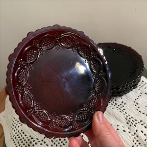 May include: A set of ruby red glass plates with a scalloped edge and ornate embossed floral design. The plates are round and appear to be vintage, with a glossy finish. A stack of similar plates is visible in the background.