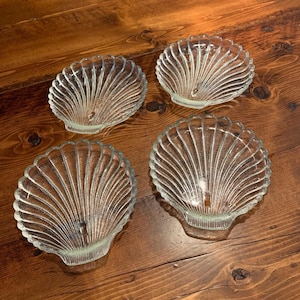 May include: Four clear glass seashell-shaped dishes. Each dish has a scalloped edge and a fan-like pattern of ridges radiating from the center. The dishes are arranged on a wooden surface.