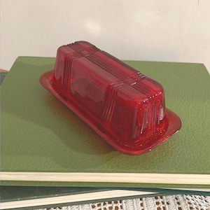 May include: A rectangular ruby red glass butter dish with a matching lid. The lid features a geometric stepped design with vertical ridges and a smooth central panel. The base has a flat rim and a recessed center to hold the lid securely in place.