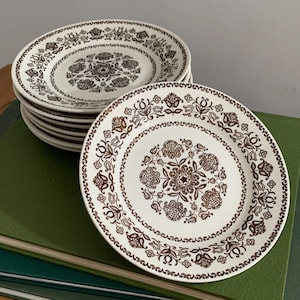 Puede incluir: Una pila de platos redondos de cerámica con una base de color crema y un patrón floral y de cachemira en marrón oscuro. El diseño incluye un motivo circular central rodeado por un borde decorativo de flores y hojas estilizadas.