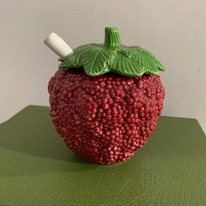 May include: A ceramic strawberry-shaped jar with a textured red surface and a removable green lid featuring molded leaf details. A small white ceramic spoon is inserted into the side of the lid, which has a central stem handle.