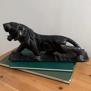 Puede incluir: Escultura de tigre de madera tallada de color marrón oscuro en pose de rugido. El tigre está sobre una base tallada y descansa sobre una pila de libros verdes. La escultura está sobre una superficie de madera.