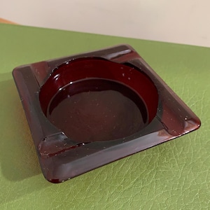 May include: A square, dark red ashtray with a circular indentation in the center. The ashtray has a glossy finish and is made of a solid material. The ashtray is sitting on a green surface.
