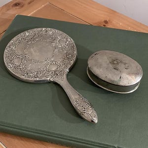 May include: An antique silver-toned hand mirror and a small oval-shaped container. The mirror has an ornate floral design around the edges and on the handle. The container has a simple, slightly tarnished surface.
