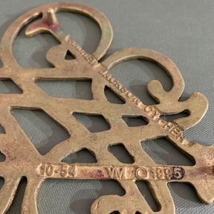 Op de afbeelding: Een vintage decoratief stuk van messing met een open ontwerp. Het stuk heeft de tekst "Andrew Jackson Cypher" en de nummers "10-54" en "1995". Het messing heeft een aangetaste, verouderde uitstraling.