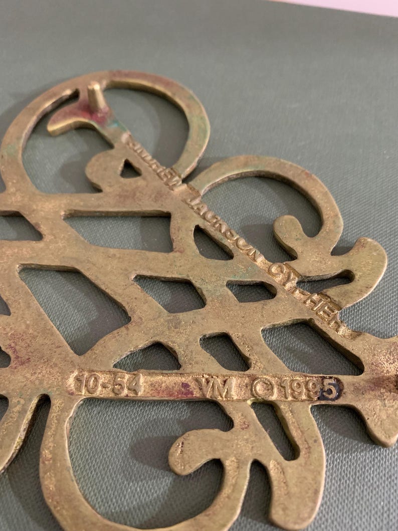 Op de afbeelding: Een vintage decoratief stuk van messing met een open ontwerp. Het stuk heeft de tekst "Andrew Jackson Cypher" en de nummers "10-54" en "1995". Het messing heeft een aangetaste, verouderde uitstraling.