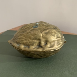 May include: A brass-coloured, walnut-shaped container with a hinged lid. The surface has a textured, wrinkled appearance, and the container is resting on a green surface. The container is likely used for storage or decoration.