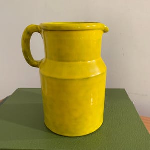 May include: A bright yellow ceramic pitcher with a glossy finish and a single curved handle. The vessel features a cylindrical body with a slightly tapered neck and a small pouring spout at the rim, showcasing subtle dark shading along the handle.
