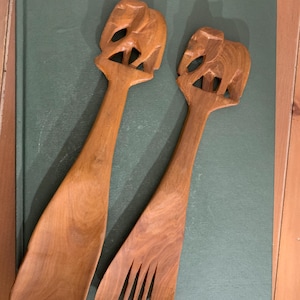 Puede incluir: Dos utensilios para ensalada de madera. Uno es una cuchara, el otro un tenedor. Ambos tienen un diseño de elefante tallado en la parte superior del mango. Los utensilios son de madera de color marrón claro y descansan sobre un libro verde.