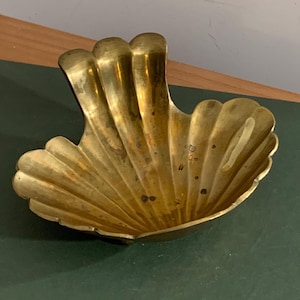 May include: A brass seashell-shaped dish with a textured surface. The dish has a scalloped edge and a curved, fan-like design. The brass has a warm, golden hue and shows signs of age.