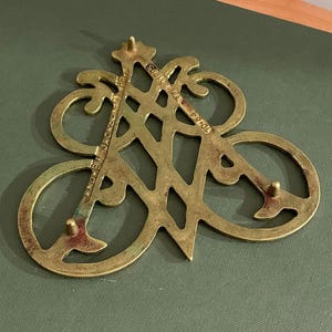 Op de afbeelding: Een vintage messing trivet met een opengewerkt ontwerp. De trivet heeft een gestileerd monogram met de tekst "ANDREW JACKSON" en "70-84". Het messing heeft een aangetaste, verouderde uitstraling, met drie kleine voetjes.