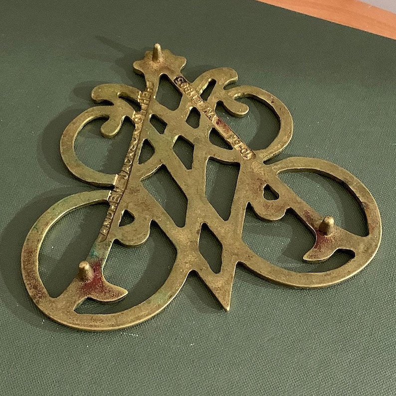 Op de afbeelding: Een vintage messing trivet met een opengewerkt ontwerp. De trivet heeft een gestileerd monogram met de tekst "ANDREW JACKSON" en "70-84". Het messing heeft een aangetaste, verouderde uitstraling, met drie kleine voetjes.
