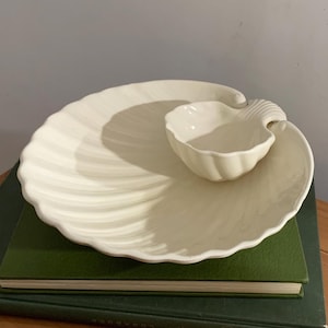 Puede incluir: Juego de fuentes de cerámica blanquecina. El juego incluye una fuente grande con forma de concha y un cuenco más pequeño con forma de concha. La fuente tiene una textura acanalada, y el cuenco se encuentra sobre la fuente.