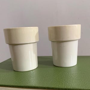 Könnte beinhalten: Zwei weiße Keramikbecher mit cremefarbenem Rand. Die Becher haben eine zylindrische Form mit leicht ausgestelltem Oberteil. Die Becher werden auf einer grünen Oberfläche präsentiert.