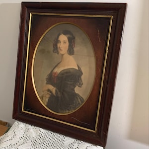 May include: A rectangular dark wood frame with a gold inner border containing an oval portrait of a person with dark hair styled in ringlets, wearing a dark off-the-shoulder garment with lace details and a red accent at the center of the neckline.