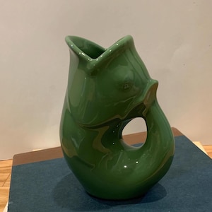 May include: A glossy, green ceramic jug shaped like a fish. The jug features a wide opening at the top, a handle formed by the fish's tail, and a circular opening in the body. This is a unique and decorative design.