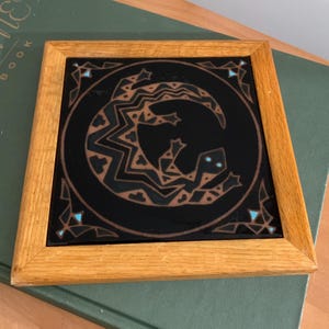 May include: Square wooden framed tile with a black background and a copper-colored lizard design. The lizard is surrounded by a circular border and accented with turquoise-colored geometric shapes. The frame is made of light-colored wood.