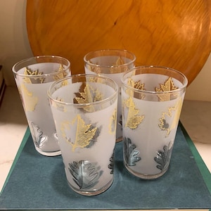 May include: Four vintage drinking glasses with a frosted finish. Each glass features a gold and silver leaf design. The glasses are cylindrical and sit on a blue-green surface, with a wooden background.