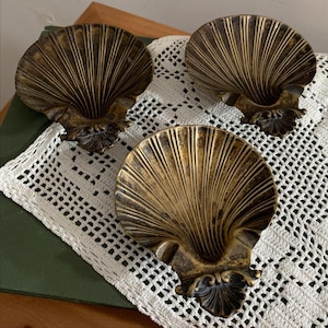 May include: Three matching brass-toned metal dishes shaped like open seashells. Each dish features a deep, fluted interior with radiating lines and a decorative, ornate hinge-like detail at the base. The metal has a dark, aged patina finish.