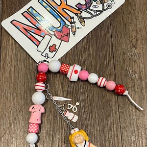 May include: A beaded nurse-themed accessory featuring a variety of pink, white, and red beads. The accessory includes a charm with a nurse figurine, a thermometer, and a medicine bottle. A card with the word "NURSE" in colorful letters is also present.