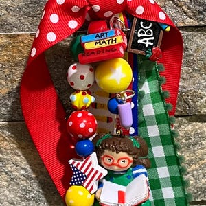 May include: A school-themed keychain with a red polka dot ribbon and green gingham ribbon. It features a miniature chalkboard with "ABC", a stack of books labeled "Art Math Reading", and a figurine of a girl reading. Colorful beads and star accents complete the design.