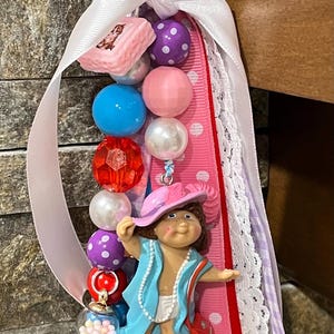 May include: A handmade keychain featuring a doll figure wearing a blue jacket and pink hat, surrounded by colorful beads and ribbons. The keychain includes a variety of beads in shades of pink, blue, and purple, and a small glass orb filled with candy.